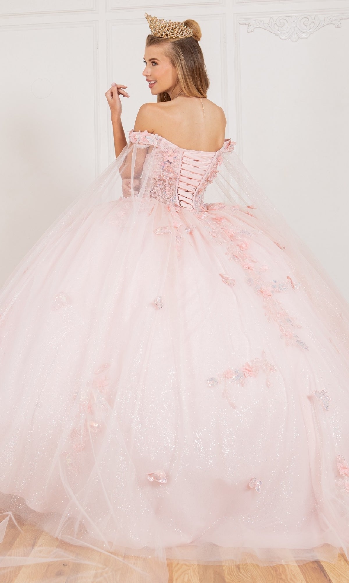 A woman in the Cinderella Couture 8075J quinceanera dress with 3D appliques, corset back, crown, and detachable sheer cape stands on a wooden floor, facing away from the camera.